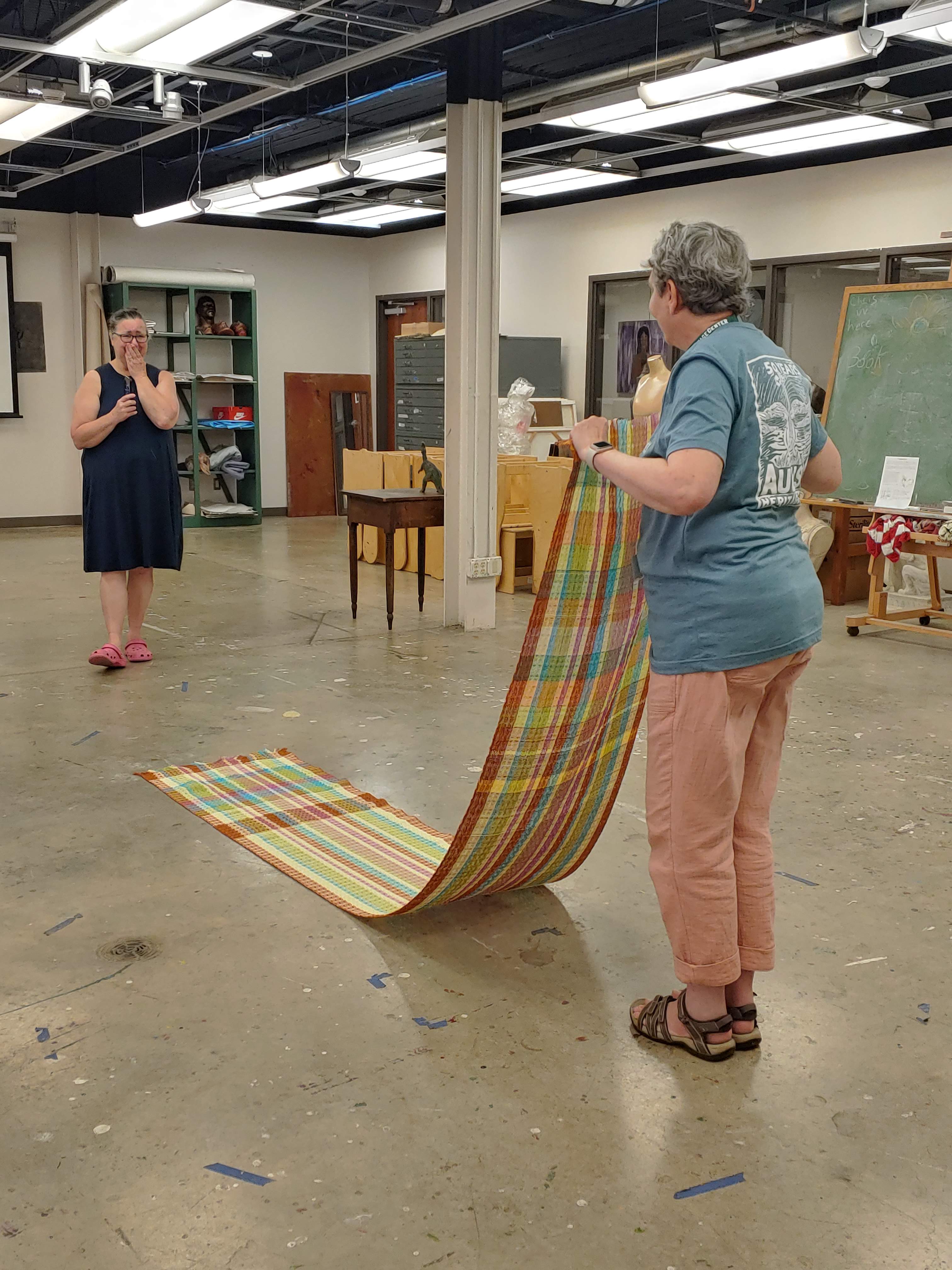 In the foreground, a woman stands with her back to the camera holding her colorful woven towels as one extended piece from the loom. She is showing her work to her teacher who stands in the background with her hand covering her mouth and smiling.
