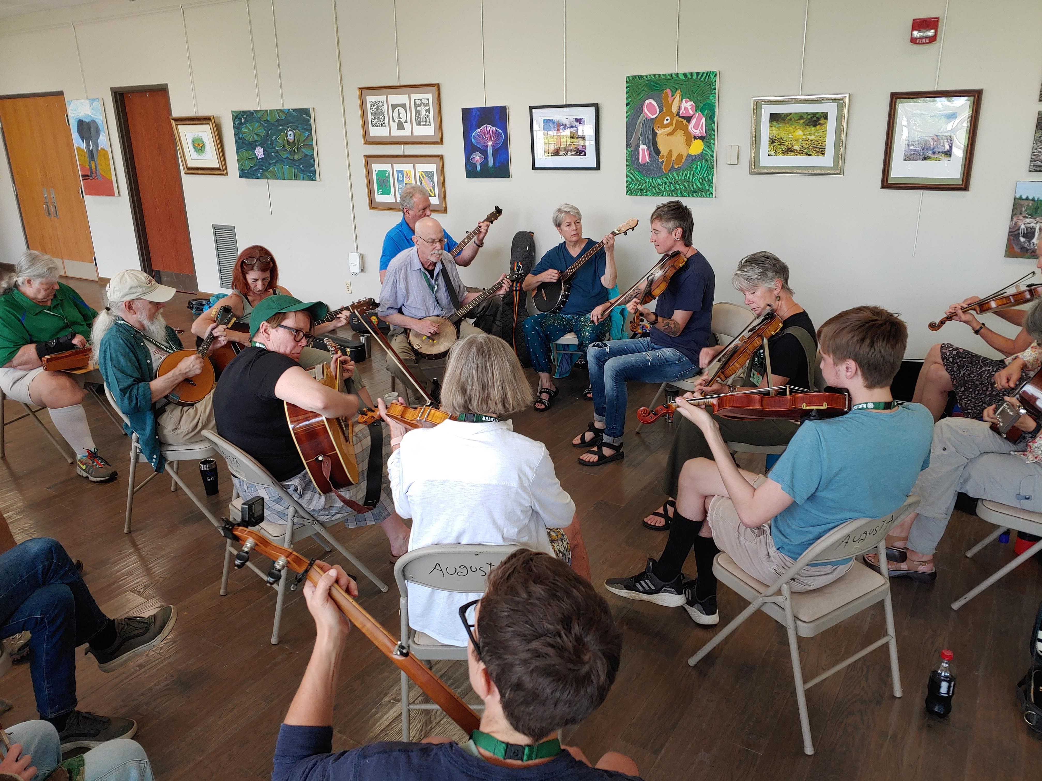 A group of about 15 people sit around a circle together playing fiddles, guitars, dulcimers, and banjos. Artwork hangs on the wall behind them.