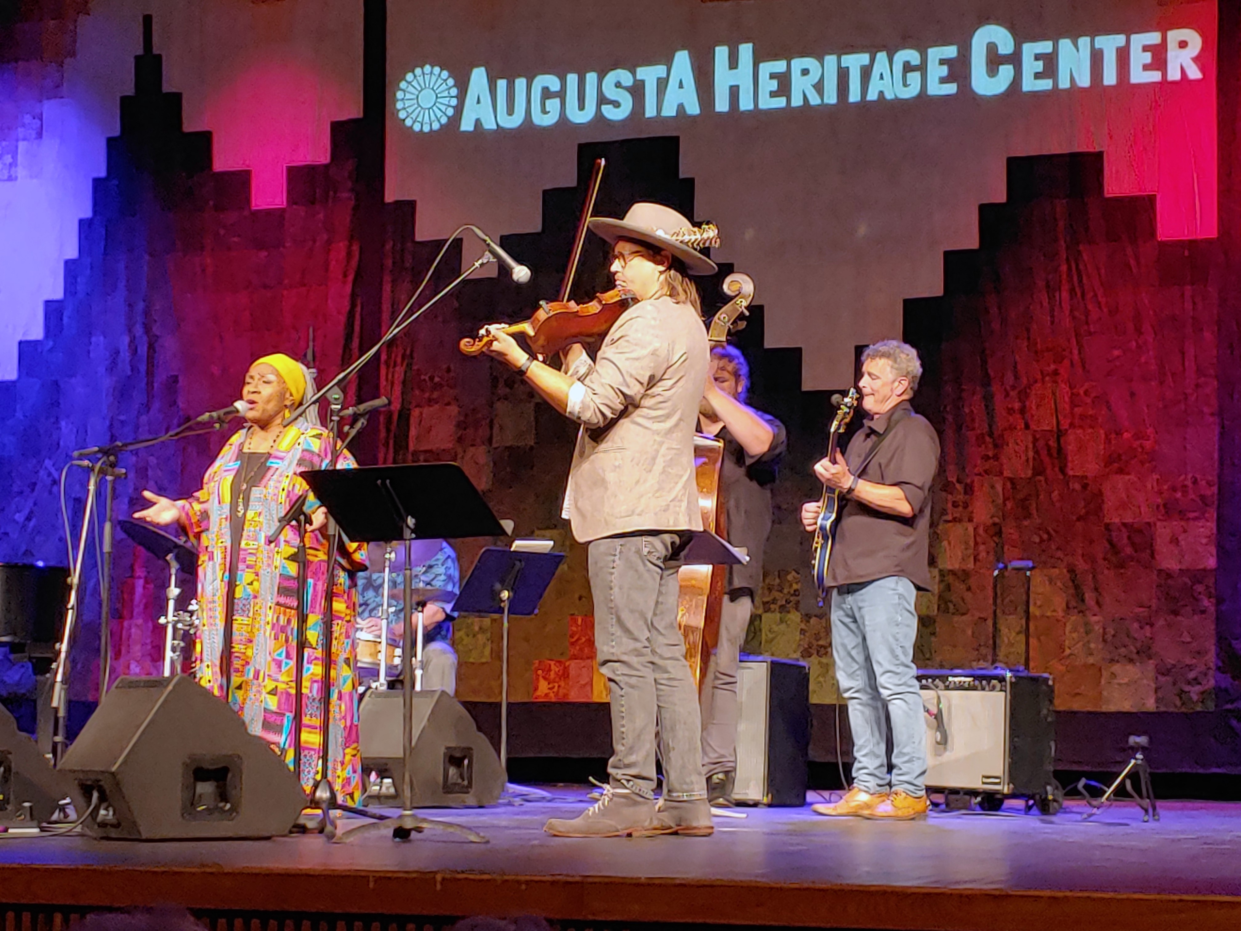A stage performance with five musicians. The backdrop is designed to look like a quilted mountain range with the Augusta Heritage Center logo projected onto it.