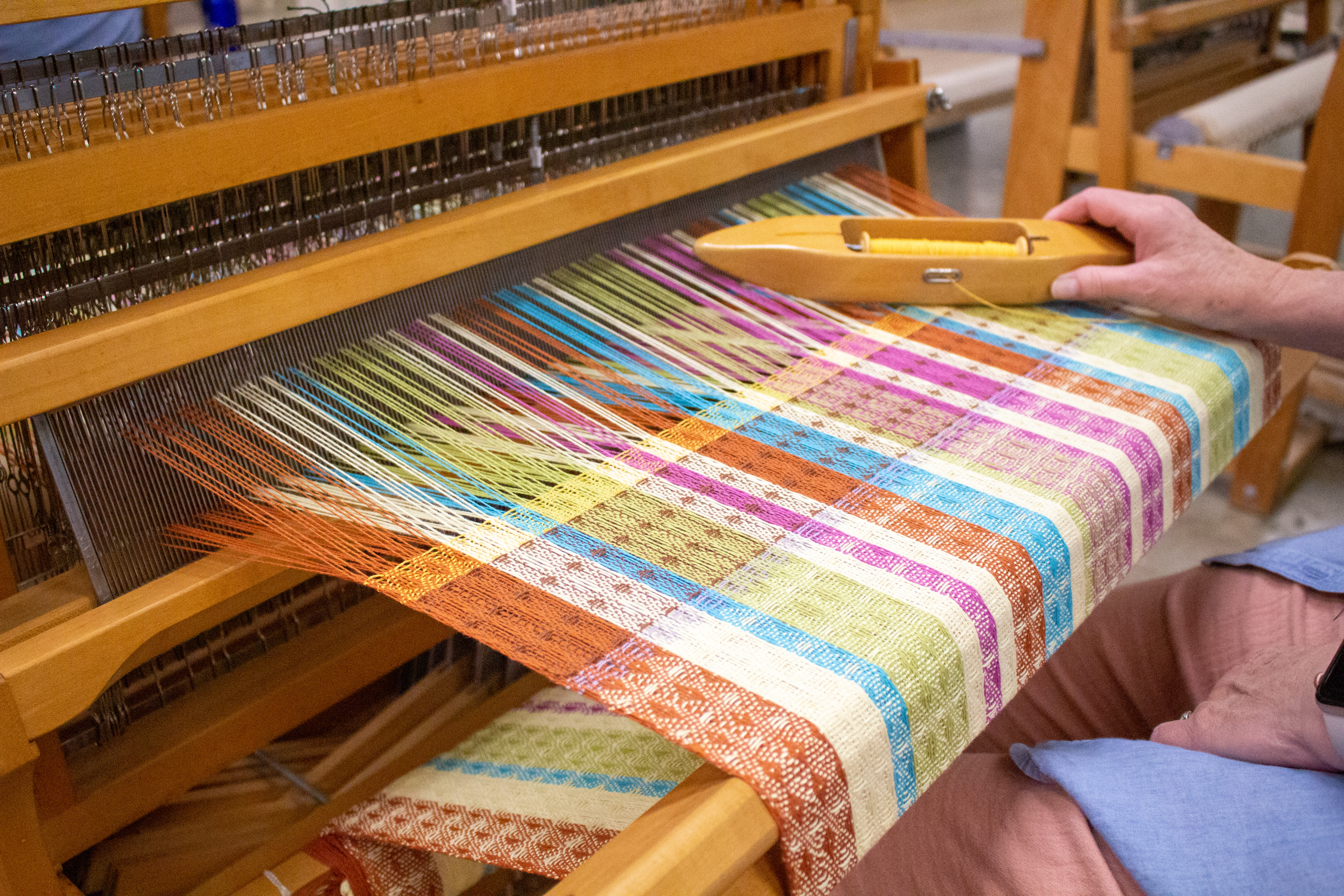 A zoomed in image of a colorful woven work in process on a loom. A hand holds a shuttle with yellow thread in the background, ready to continue weaving.