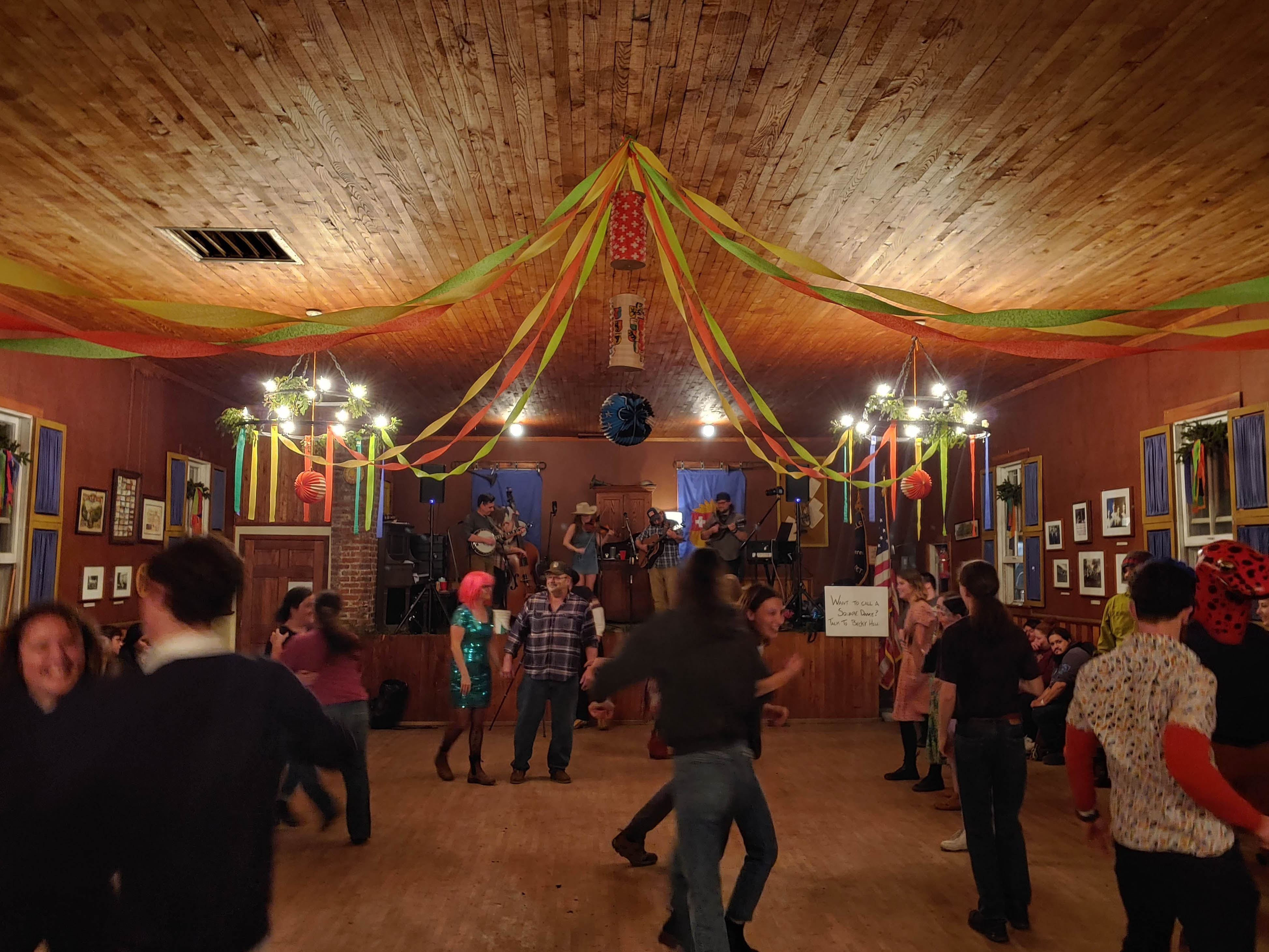 Inside a dance hall, colorful streamers overhead. A string band plays in the background on a stage for dancers in the foreground.