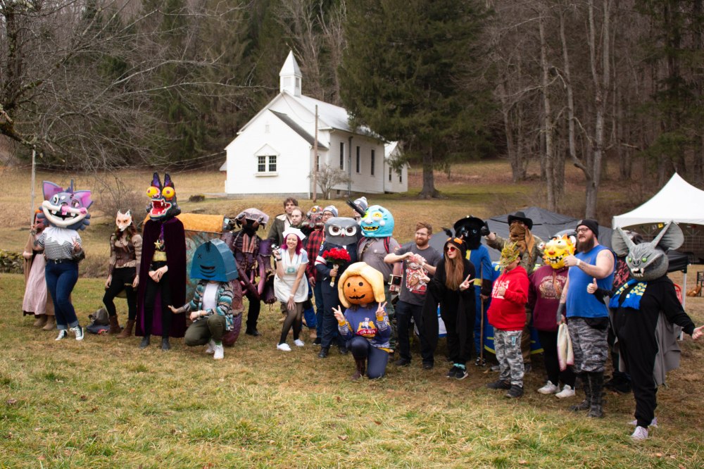 Foreground: Masked people stand for a photo outside. Background: a small white church and forest.