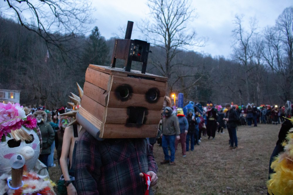 A crowd of masked people stand outside in a large circle during the mask contest. Foreground: A person with a wooden box as a mask with two holes cut for eyes and a square for a mouth, with what looks like a wood stove for a hat.