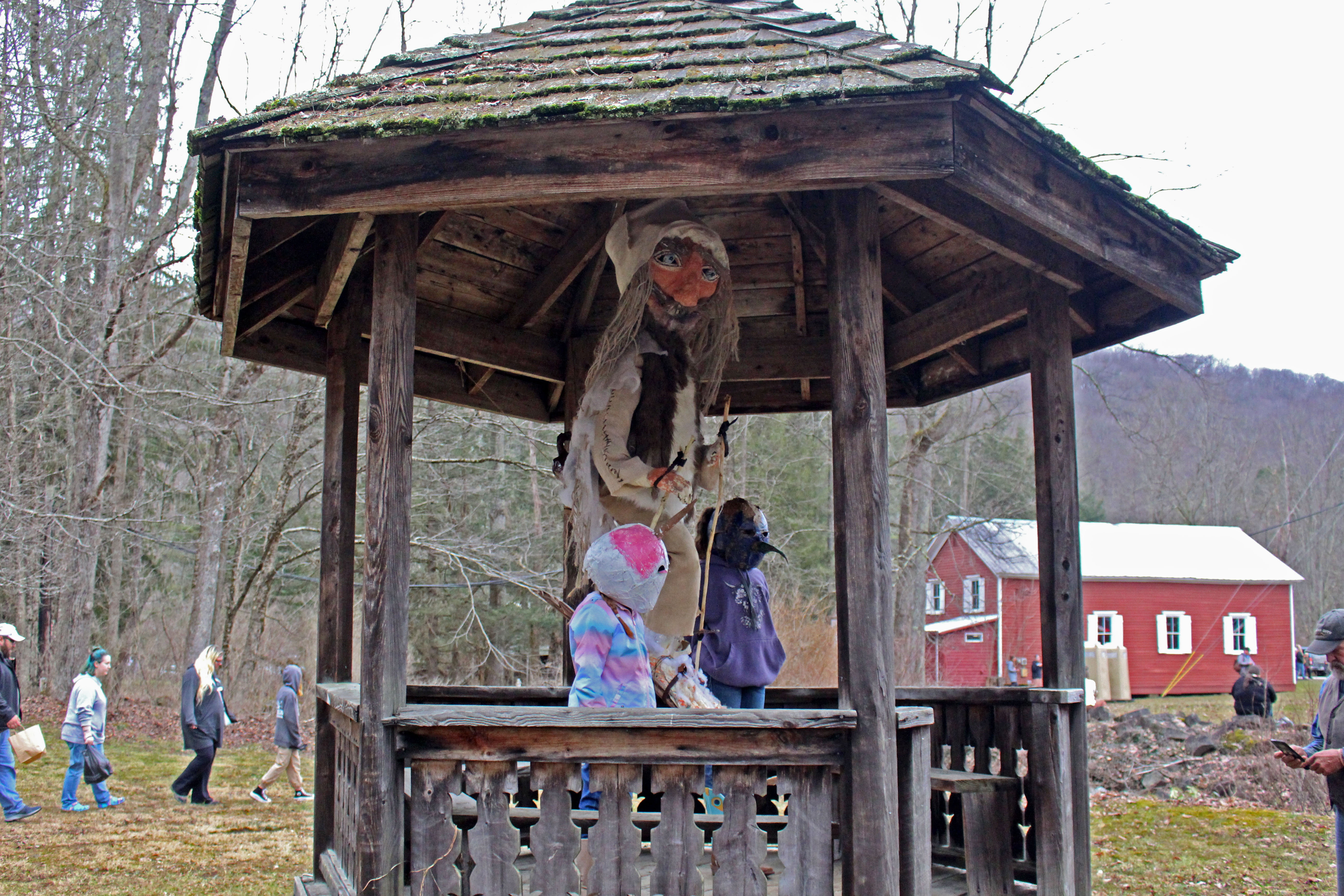 An effigy of Old Man Winter made out of fiber materials hangs in a gazebo outdoors as two children stand beside it wearing round colorful masks.