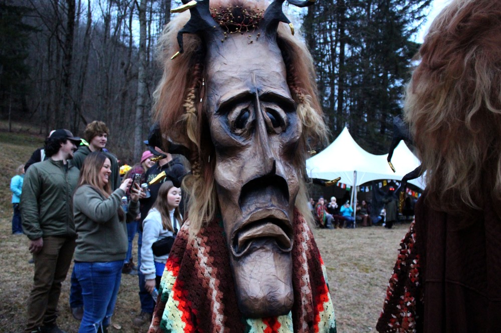 A crowd of people stand outside during the mask contest. Foreground: a person wearing an enormous mask of a sad face with horns and a crocheted afghan blanket over their shoulders.