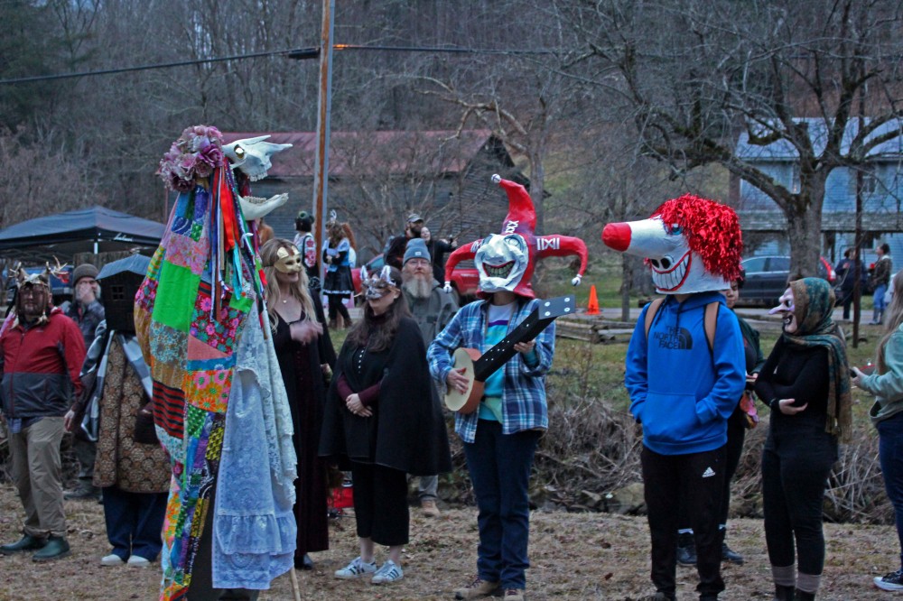 A crowd of people stand outside during the mask contest. One with a skull and a colorful quilt-style dress, another that looks like a clown with a banjo, and one that looks like a clown.