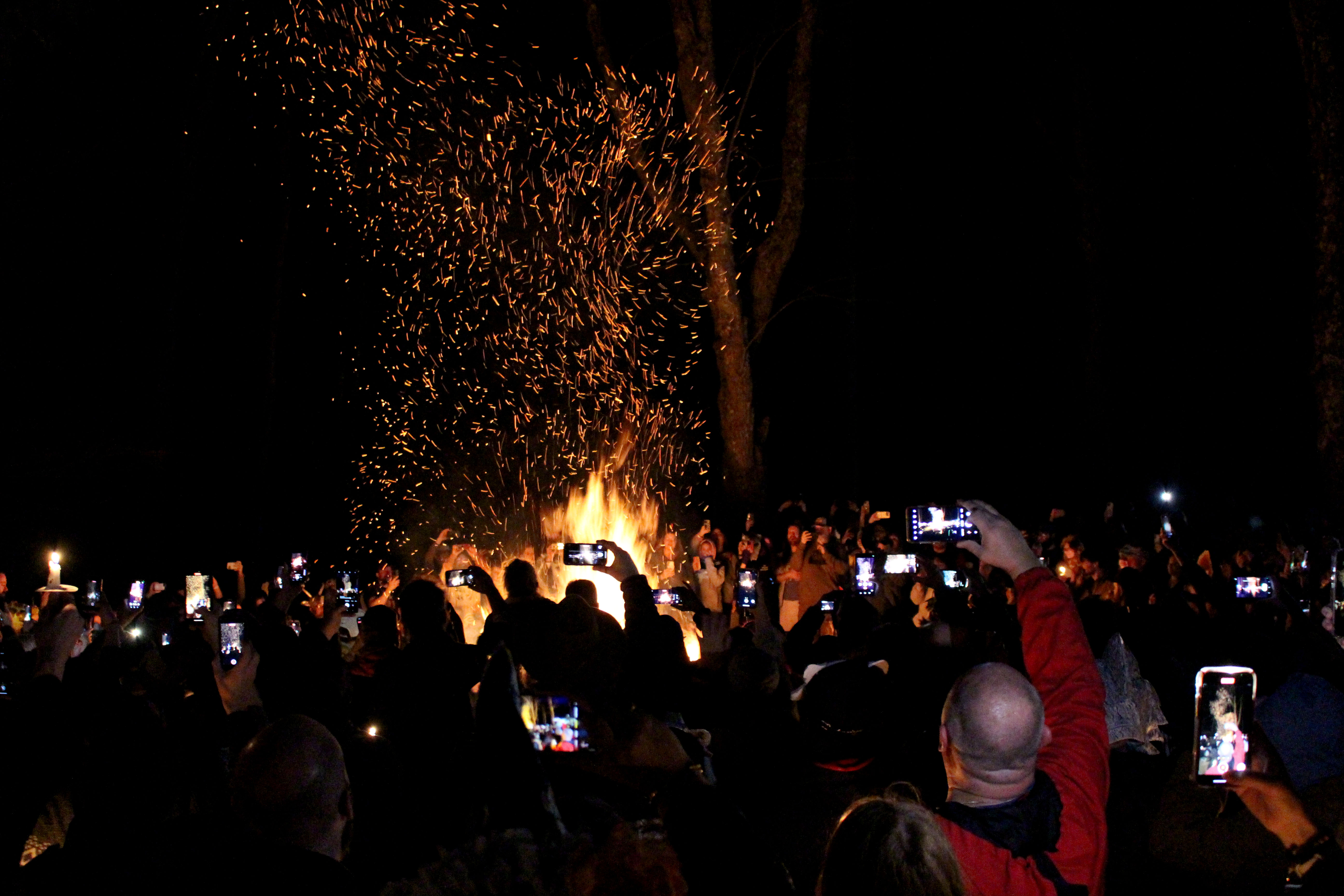 At night, a crowd surrounds a bonfire and takes photos of the flames.
